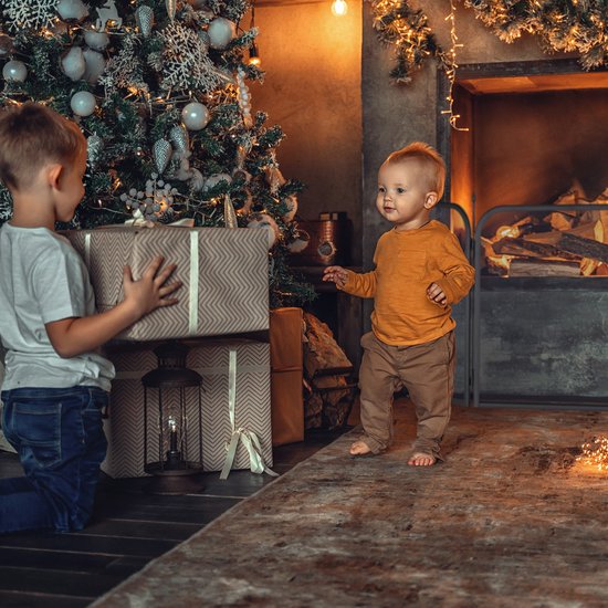 Kinderen bij een kerstboom met cadeaus, met een open haard op de achtergrond.