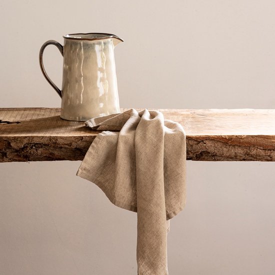 Crèmekleurige kan met bruine rand, rustend op een houten plank met beige linnen servet.