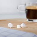 Drie witte Krups reinigingstabletten, één met de letter K, op een houten plank, met een kopje koffie op de achtergrond.