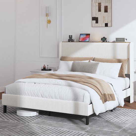 Lit rembourré crème avec tête de lit côtelée. Lit fait avec literie blanche et marron. Une tablette et un téléphone sur la tête de lit.