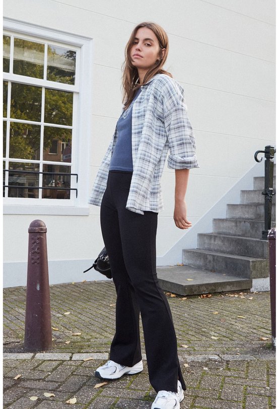 Vrouw met zwarte wijduitlopende legging, grijs geruite blouse en witte sneakers, met de hand op de enkel.