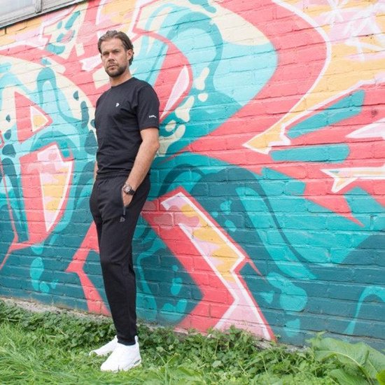 Man in zwarte joggingbroek en zwart T-shirt, staand voor een muurschildering.