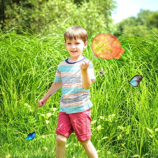 Een jongen in een gestreepte top en rode korte broek lacht en houdt een oranje schepnet vast in het hoge gras met vlinders.