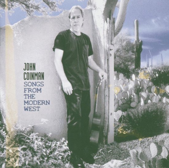 John Coinman staat met een gitaar in een woestijn met cactussen.