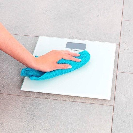 Une main avec un chiffon bleu nettoyant une balance personnelle numérique blanche.
