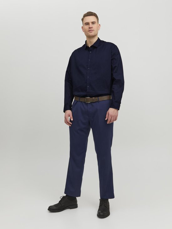 Man staat met handen in zakken, draagt een donkerblauw overhemd met lange mouwen en een marineblauwe broek met bruine riem.