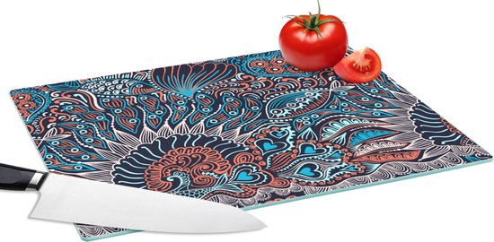 Planche à découper en verre avec un motif abstrait de poissons et de fleurs corail et turquoise, avec un couteau et une tomate.