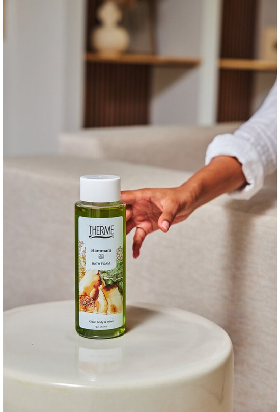 Groene fles met Hammam Badschuim van Therme op een bijzettafeltje. Een hand reikt ernaartoe. De fles heeft gouden patronen en bruine/groene illustraties.