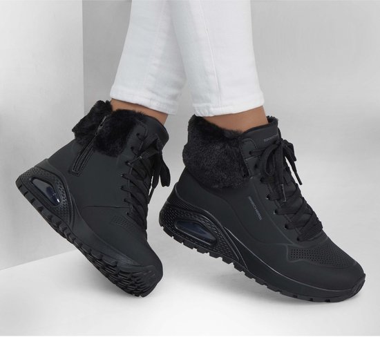 Model draagt zwarte Skechers Uno Rugged veterboots met witte broek, met bontkraag en Air-zool.