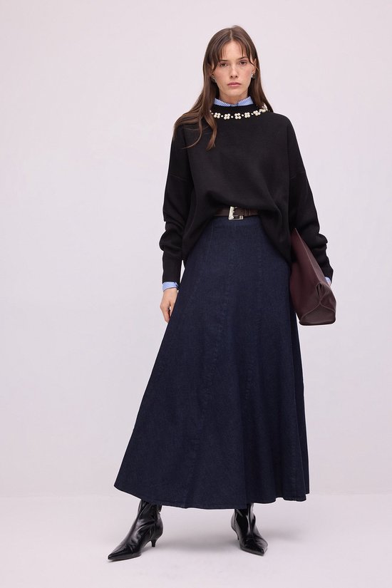 Model draagt een zwarte trui met parelversiering en een donkerblauwe denim rok, afgewerkt met een bruine riem.