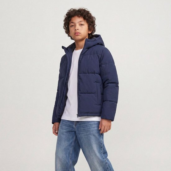 Jongen met krullend haar in een donkerblauwe pufferjas met capuchon, wit T-shirt en jeans.