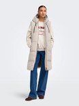 Model draagt een lange beige bodywarmer met capuchon over een crème sweater met 'Paris Social Club' opdruk en een spijkerbroek.