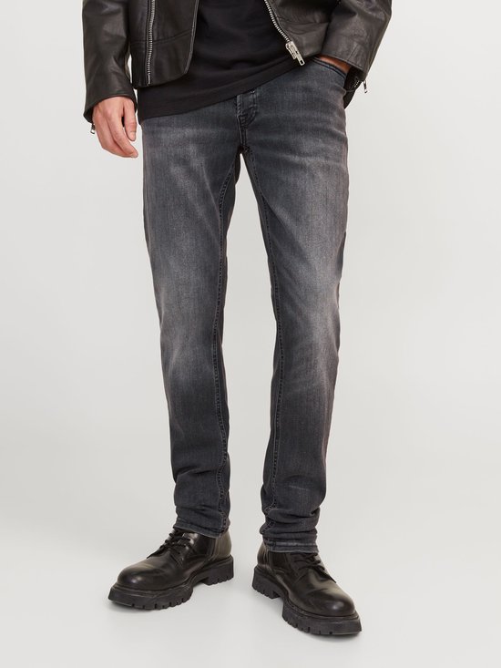 Man zittend op een krukje, met zwart T-shirt, leren jack en grijze jeans.