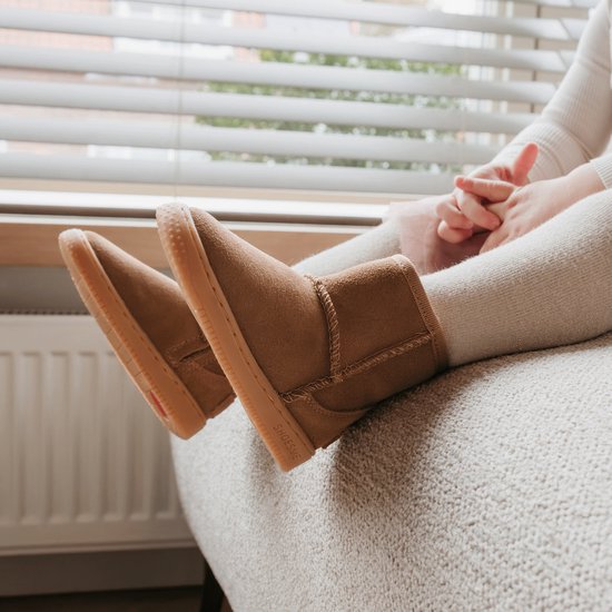 Voeten van een kind in bruine suède laarsjes met witte zolen, zittend op een stoel.