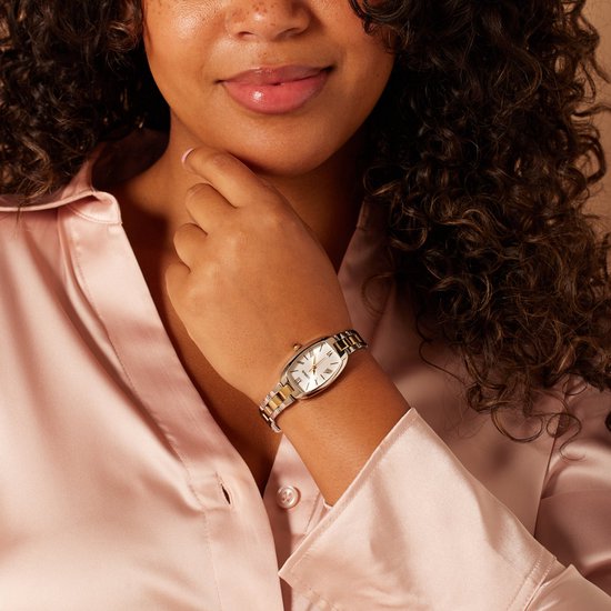 Gouden en zilverkleurig horloge met witte wijzerplaat gedragen door een vrouw in een roze satijnen blouse.