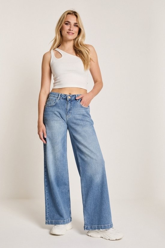 Model in een witte top met één schouder en wijde, lichtblauwe jeans.
