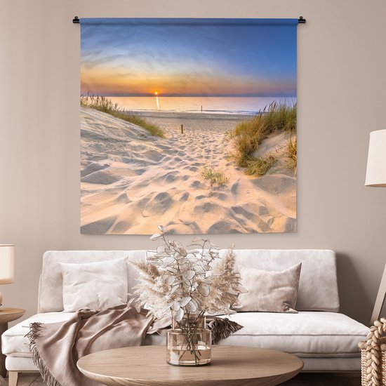 Wandkleed met duinlandschap bij zonsondergang, hangend in een woonkamer boven een beige bank met decoratie.
