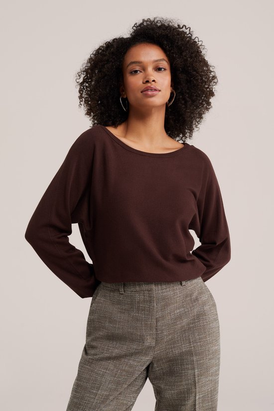 Model met een bruine trui met boothals, handen achter de rug en een geruite broek.
