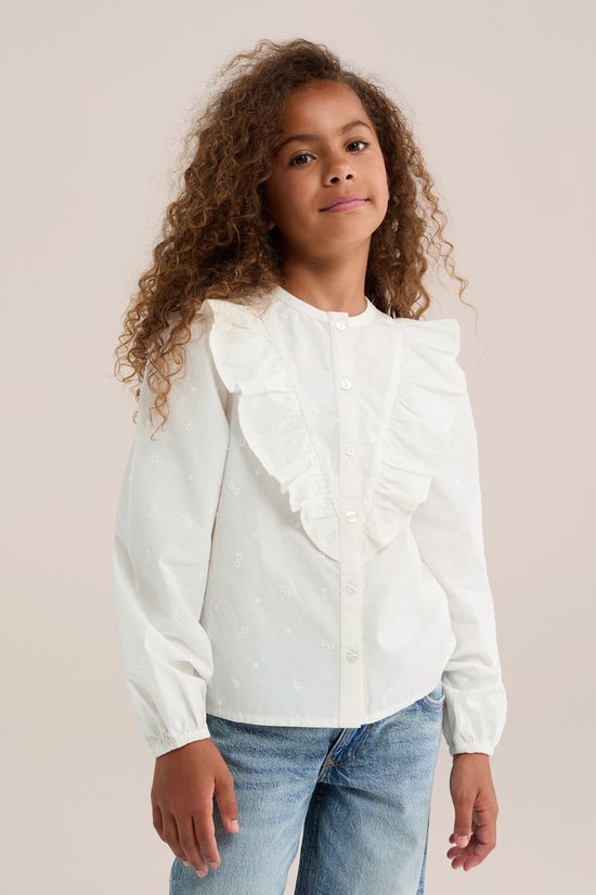 Meisje met krullend haar draagt een witte blouse met ruches en geborduurde bloemetjes, gecombineerd met een spijkerbroek.