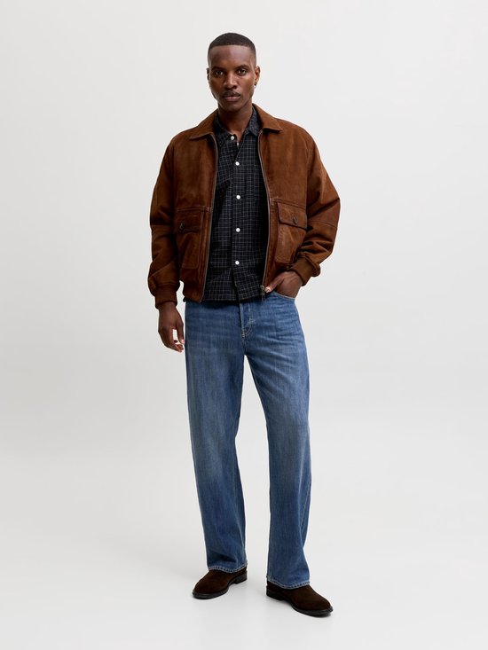 Man met bruine jas, geruite blouse en wijde blauwe jeans, staand.