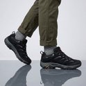 Man met grijze sokken en broekspijpen opgerold, draagt zwarte Merrell wandelschoenen.