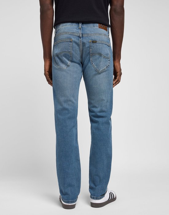 Man met de rug naar ons toe, draagt een lichtblauwe spijkerbroek, een zwart T-shirt en witte sneakers met strepen.