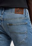 Detail van de achterkant van een lichtblauwe spijkerbroek met een bruin lederen Lee-patch en een klein Lee-logo.