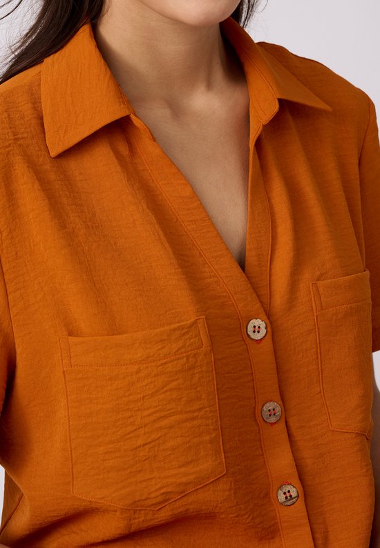 Detail van oranje blouse met korte mouwen, kraag, twee borstzakjes en houten knopen.