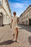 Model draagt een beige jurk en witte schoenen op een straat.