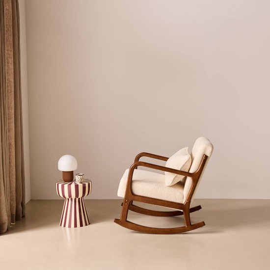 Schommelstoel met walnootkleurig houten frame, crèmekleurige bouclé bekleding en een bijpassend kussen. Naast de stoel staat een gestreepte bijzettafel met lamp.
