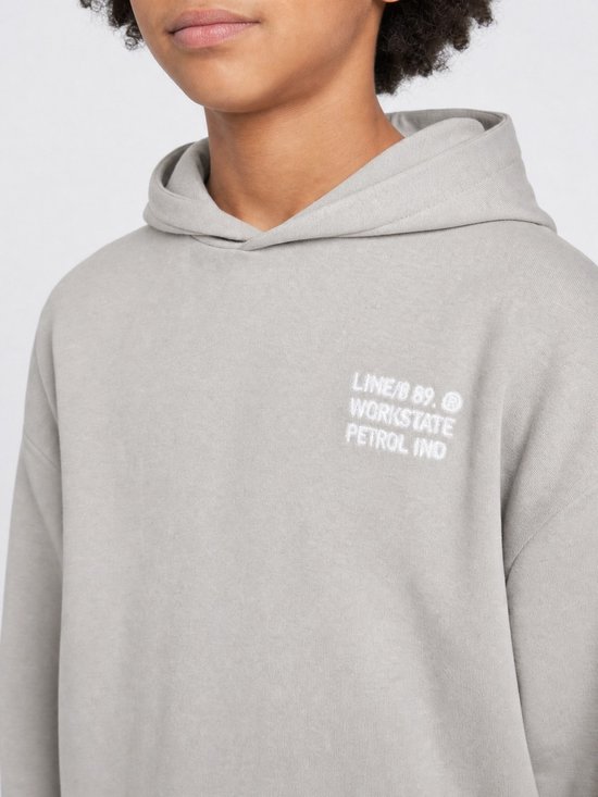 Close-up van een jongen in een lichtgrijze hoodie met kleine witte tekst op de borst.