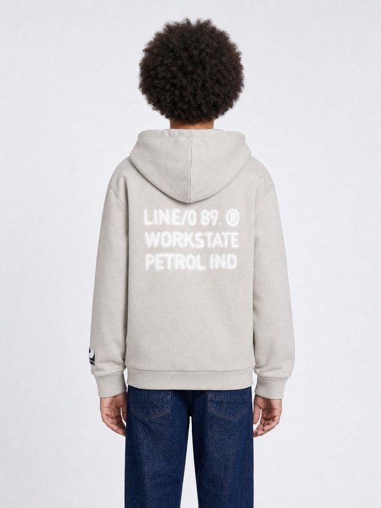 Jongen draagt lichtgrijze hoodie met tekst op de rug en donkere jeans. De capuchon is op.