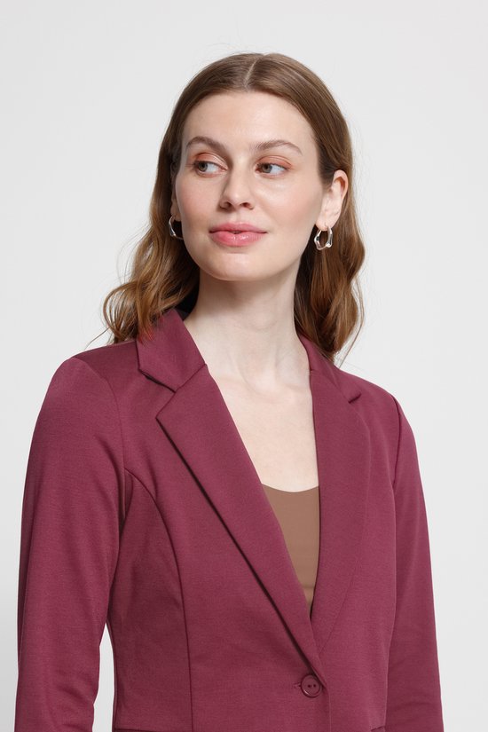 Vrouw met lang golvend bruin haar, draagt een bordeauxrode blazer en oorringen. Kijkt naar rechts.