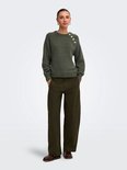 Vrouw in donkergroene trui met knopen op de schouder, staand met broek en schoenen.