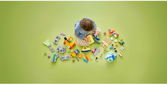 Kind met LEGO Duplo trein, station, voertuigen en accessoires op groene achtergrond.