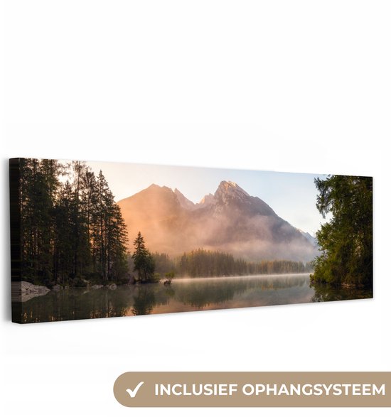 Panoramisch schilderij van een mistig berglandschap bij zonsopkomst, met bomen en reflectie in het meer. Inclusief ophangsysteem.
