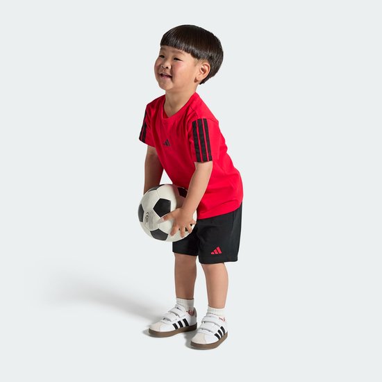 Jongetje in rood shirt met zwarte strepen en zwarte shorts, met witte sneakers en een voetbal.