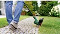 Persoon in spijkerbroek en grijze schoenen maait gras met groene grastrimmer op een betegeld pad.