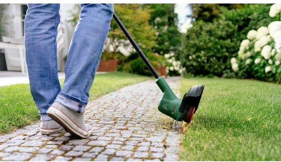 Persoon in spijkerbroek en grijze schoenen maait gras met groene grastrimmer op een betegeld pad.