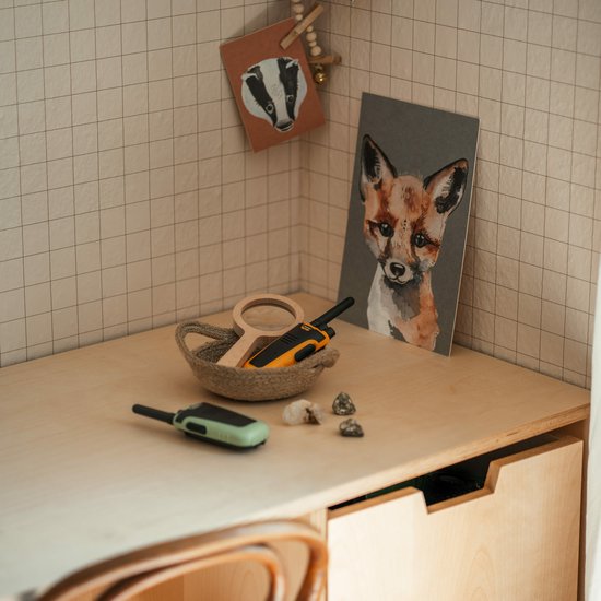 Groene en oranje Kidywolf KIDYTALK walkietalkies op een houten bureau, met tekeningen aan de muur.