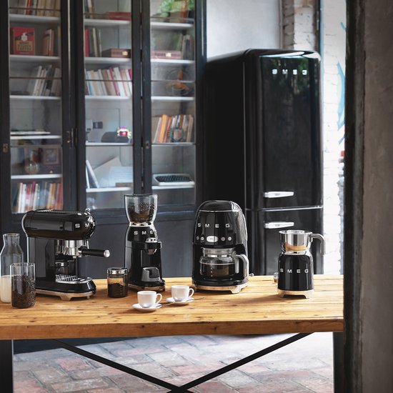 Zwarte SMEG keukenapparaten waaronder een koffiezetapparaat, melkopschuimer en koelkast op een houten tafel.