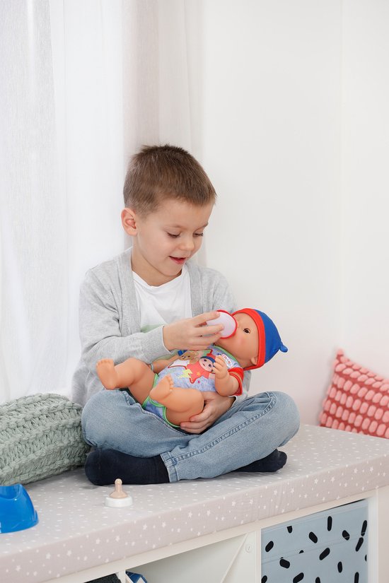 Jongen geeft BABY born pop flesje. Zittend op een bankje met witte lakens.