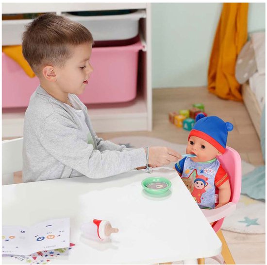 Jongen voert BABY born Jonas pop, zittend in een roze stoel, met een lepel uit een schaaltje.