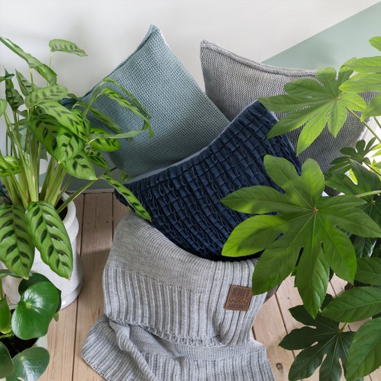 Een stapel gebreide kussens in grijs, blauw en een donkerblauw kussen met textuur, omringd door groene planten.