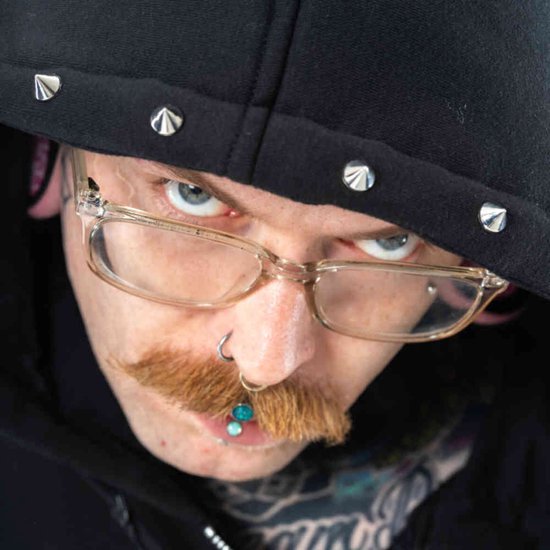 Close-up van een persoon met bril, neusring en doordringende blik, draagt een zwarte hoodie met zilveren studs op de capuchon.