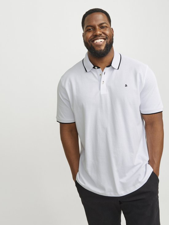 Man met baard draagt lichtgrijze poloshirt met zwarte biezen en zwarte broek, glimlachend.