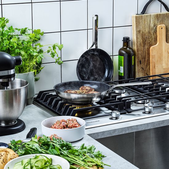 Man met witte T-shirt kookt op gasfornuis met verschillende pannen, ingrediënten en keukenapparatuur.