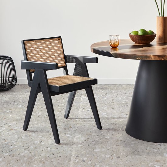 Zwarte stoel met rieten zitting en rugleuning in een eetkamer met terrazzo vloer en houten tafel.