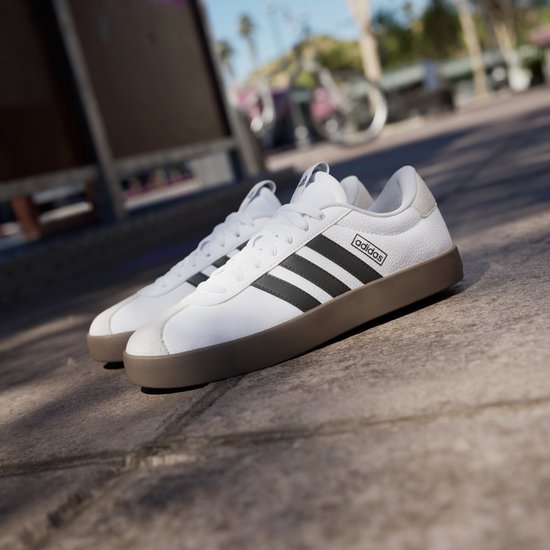 Witte Adidas VL Court 3.0 sneakers met zwarte strepen en bruine zool, op een buitenlocatie.