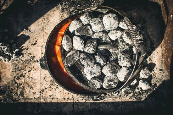 Gietijzeren pan met oranje stoofpot, bedekt met kolen, gezien van bovenaf.